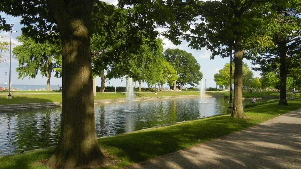 A tree-lined lakefront park path with a fountain and open water beyond