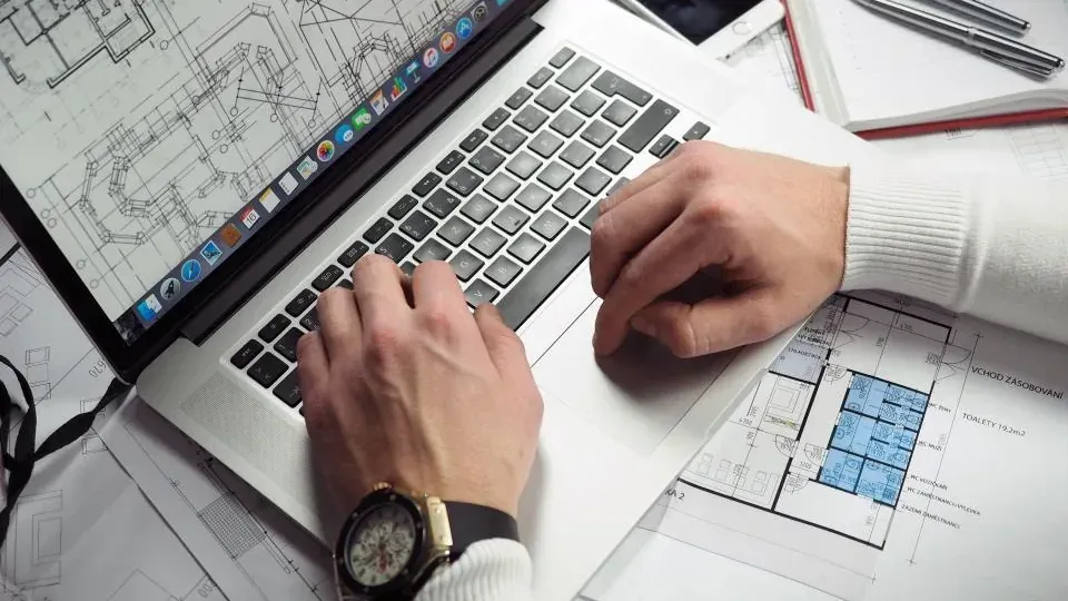 Hands on a laptop keyboard beside printed floor plans, a phone and pens on a white desk