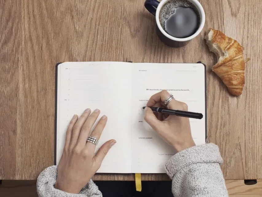 A person writing notes in an open notebook on a wooden desk beside a coffee cup