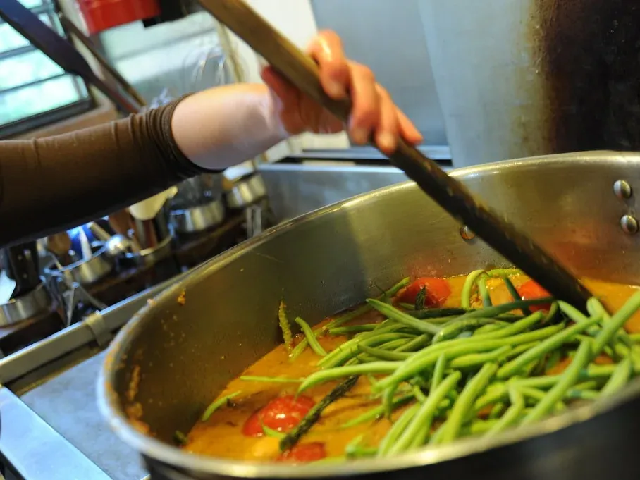 A chef stirring vegetables in a steel pan on a busy commercial kitchen range