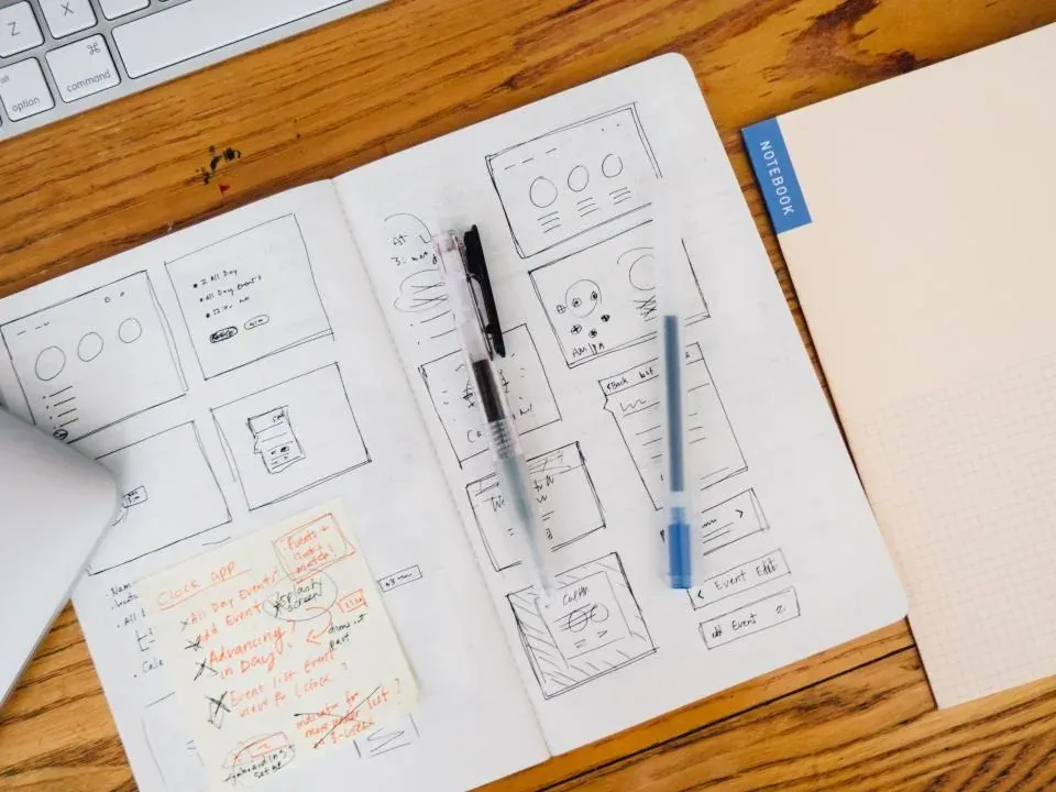 Hand-drawn interface wireframe sketches on paper beside a pen on a wooden desk